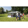 Wohnmobil mit ausgefahrener Markise auf einer Wiese; vier Personen stehen neben einem Kugelgrill; im Hintergrund Bäume und Campingstühle