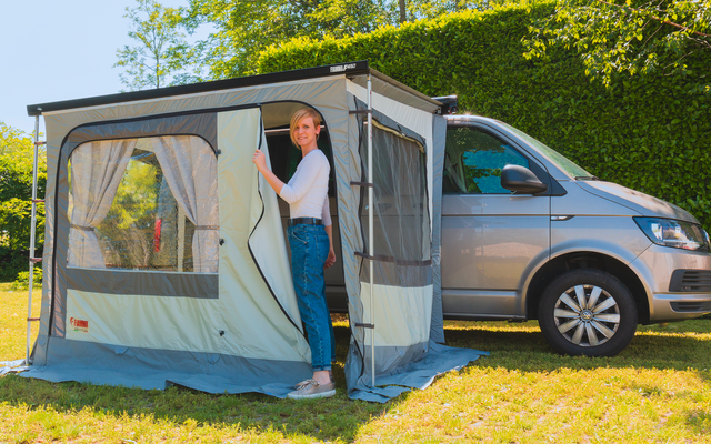 Persoon staat in een geopende voortent bevestigd aan een grijze camperbus op gras