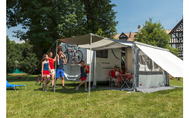 Wohnwagen mit Markise auf einer Wiese, davor zwei Erwachsene und zwei Kinder, Fahrrad am Wohnwagen, Tisch und Stühle unter der Markise, Fachwerkhäuser, Bäume und blauer Himmel