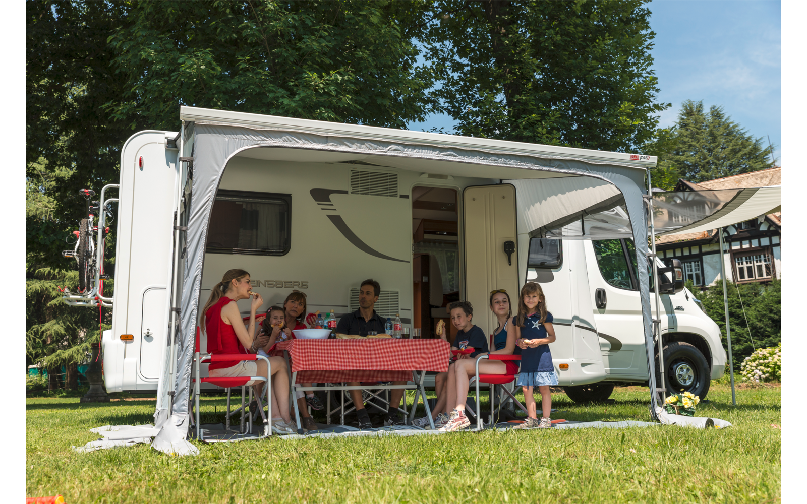 Campingbus mit ausgefahrener Markise auf einer Wiese, vor dem Bus ein Tisch mit rotem Tuch und mehrere Personen darunter, Bäume und ein Gebäude im Hintergrund