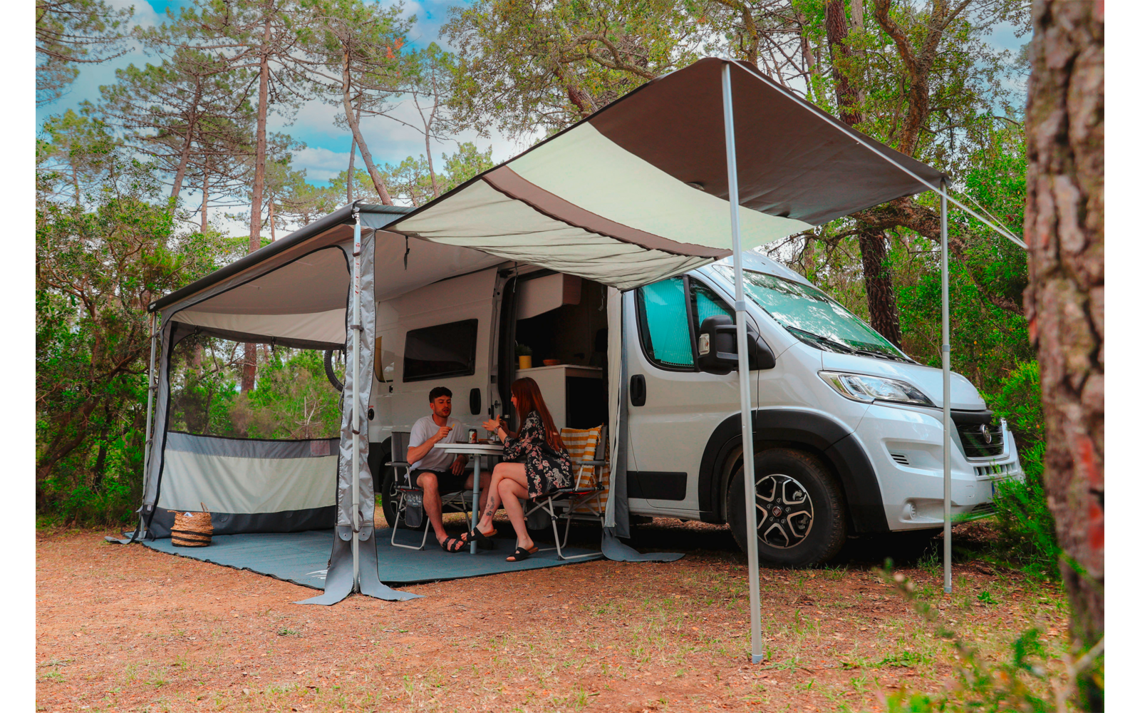 Campingbus mit ausgeklapptem Sonnensegel unter Bäumen; vor dem Bus sitzen zwei Personen an einem kleinen Tisch mit Tassen