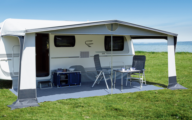 Weißer Wohnwagen mit ausgefahrener Markise, zwei Campingstühle, klappbarer Tisch, Kühlbox und Meer im Hintergrund