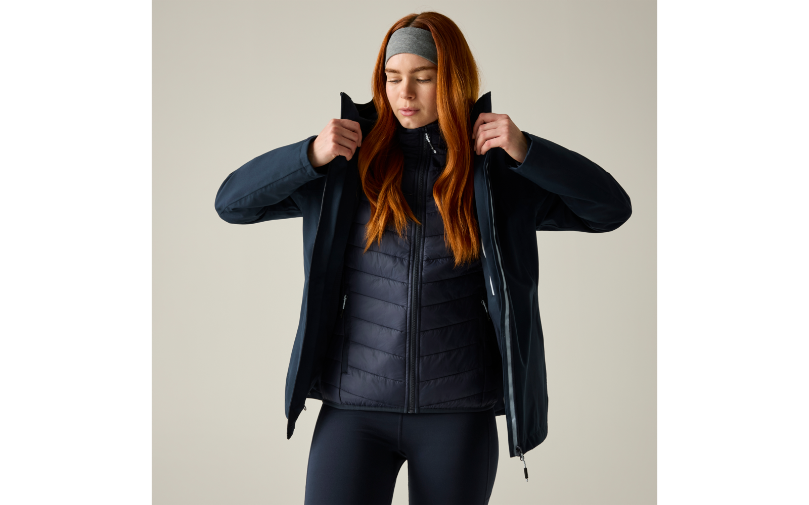 Person mit langem roten Haar und Stirnband zieht Außenjacke über Steppjacke an, Augen geschlossen, heller Hintergrund