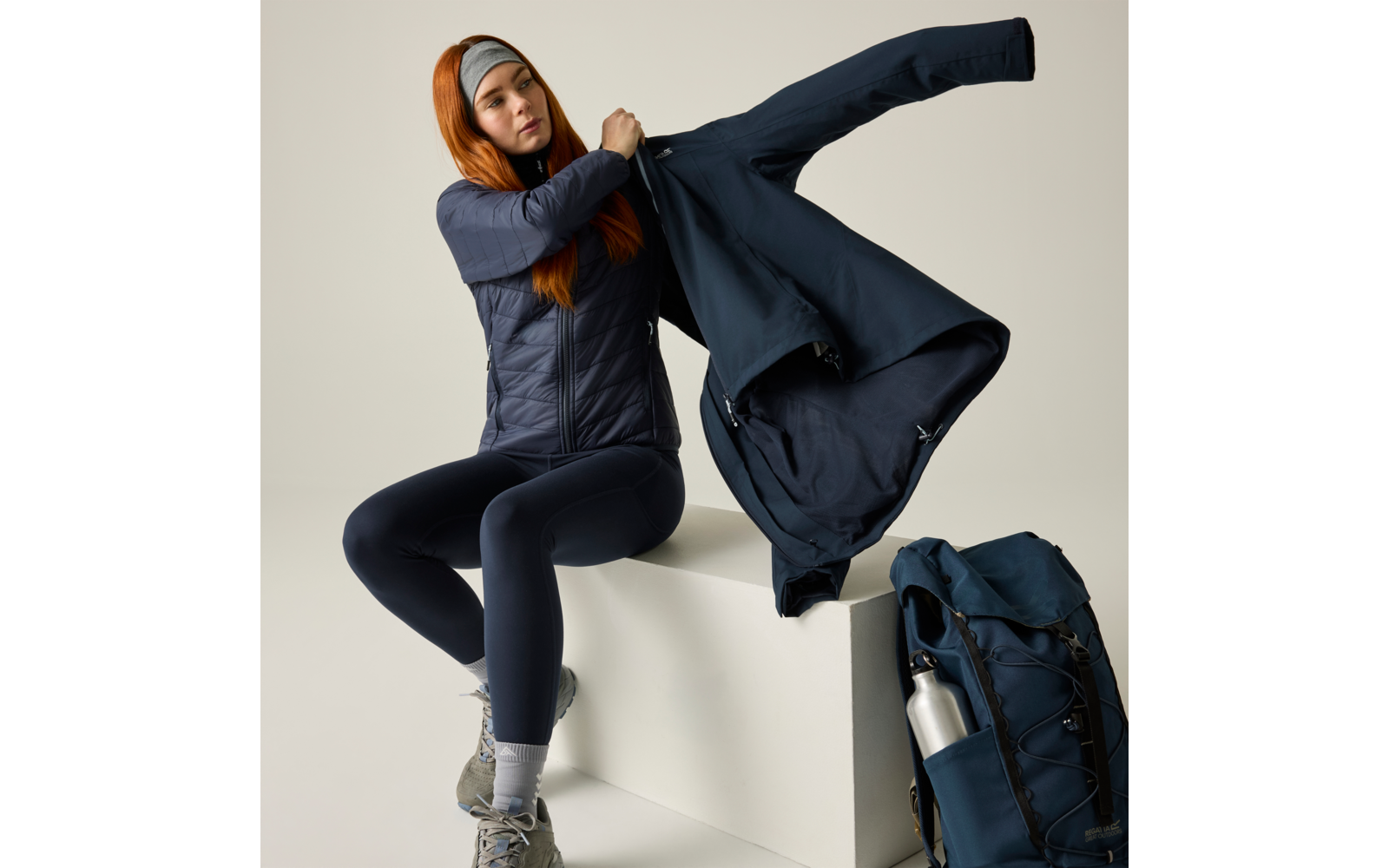 Frau mit roten Haaren und grauem Stirnband sitzt auf weißem Podest und zieht eine dunkle Jacke an; sie trägt eine gesteppte dunkelblaue Weste, dunkelblaue Leggings, graue Socken und Wanderschuhe. Neben dem Podest steht ein dunkelblauer Rucksack mit e