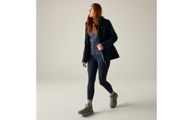 Person mit langen roten Haaren trägt dunkelblaue Outdoorjacke, graue gesteppte Weste, dunkelblaue Leggings, graue Wanderschuhe und Stirnband, geht nach rechts vor beigefarbenem Hintergrund