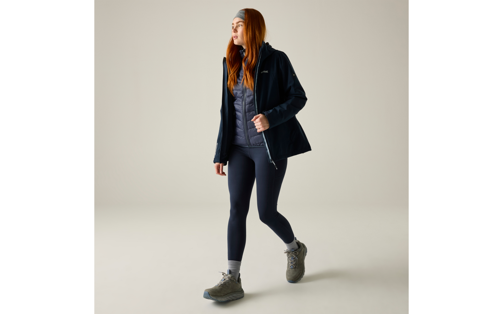Person mit langen roten Haaren trägt dunkelblaue Outdoorjacke, graue gesteppte Weste, dunkelblaue Leggings, graue Wanderschuhe und Stirnband, geht nach rechts vor beigefarbenem Hintergrund