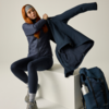 Frau mit roten Haaren und grauem Stirnband sitzt auf weißem Podest und zieht eine dunkle Jacke an; sie trägt eine gesteppte dunkelblaue Weste, dunkelblaue Leggings, graue Socken und Wanderschuhe. Neben dem Podest steht ein dunkelblauer Rucksack mit e