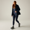 Person mit langen roten Haaren trägt dunkelblaue Outdoorjacke, graue gesteppte Weste, dunkelblaue Leggings, graue Wanderschuhe und Stirnband, geht nach rechts vor beigefarbenem Hintergrund