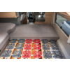 Interieur van een camper met een matras; eronder rode en grijze vierkante kunststofplaten op houten latten; raam rechts, zitkussens links, doorgang naar de cabine achteraan