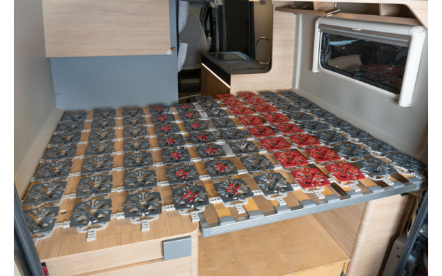 Interieur van een camper met een houten bedplatform bedekt met rijen vierkante metalen veermodules, meestal grijs met enkele rode; een raam aan de rechterkant en licht houten bekleding