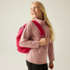 Fille aux cheveux blonds portant un sac à dos rouge sur le dos, pull rose et pantalon noir, regardant en haut à droite sur un fond clair