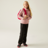 Fille blonde debout devant un fond clair, portant un pull rose, un pantalon noir et un sac à dos rouge