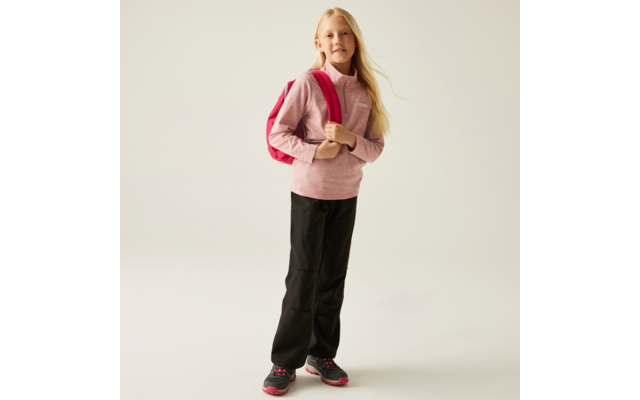 Fille blonde debout devant un fond clair, portant un pull rose, un pantalon noir et un sac à dos rouge