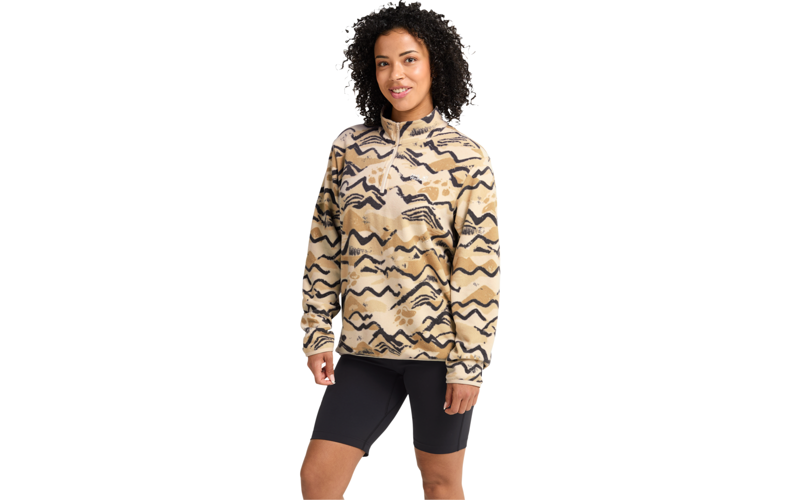 Person mit lockigem dunklem Haar, trägt beige Pullover mit kurzem Reißverschluss und abstraktem Muster sowie schwarze Radlerhosen, steht vor hellem Hintergrund