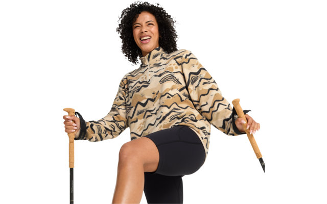 Person mit lockigen Haaren, trägt beige-schwarze Fleecejacke mit wellenförmigem Muster und schwarze Radlerhose, hält zwei Wanderstöcke mit Korkgriffen, ein Bein angehoben, lächelnd