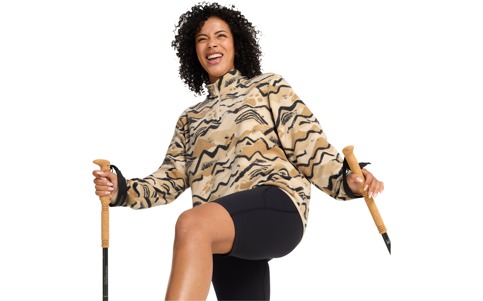 Person mit lockigen Haaren, trägt beige-schwarze Fleecejacke mit wellenförmigem Muster und schwarze Radlerhose, hält zwei Wanderstöcke mit Korkgriffen, ein Bein angehoben, lächelnd