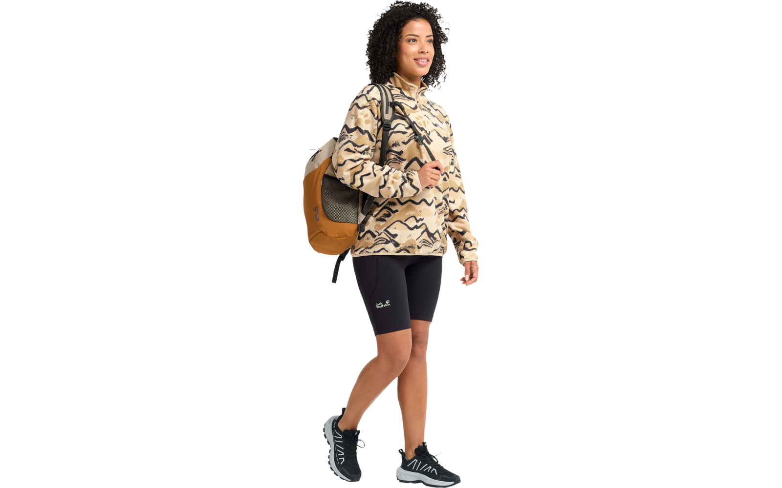 Person geht nach rechts, trägt beige-schwarz gemusterten Kapuzenpullover, schwarze Radshorts, schwarze Sportschuhe, senffarbenen Rucksack und hat dunkle, lockige Haare