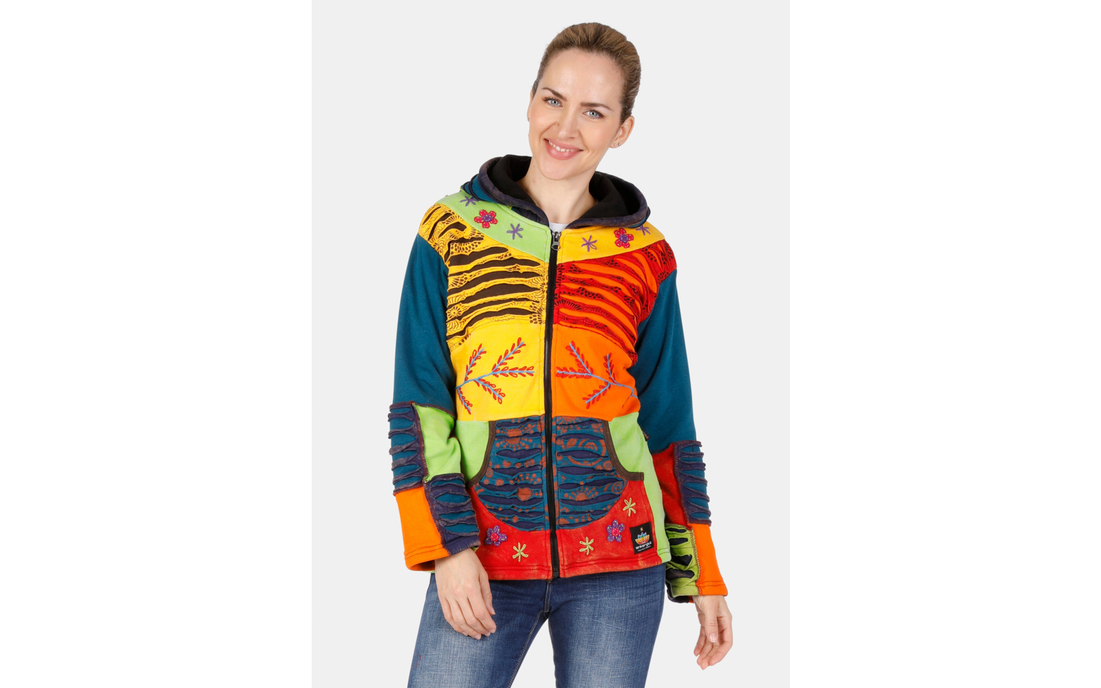 Person vor weißem Hintergrund trägt bunte, gemusterte Kapuzenjacke mit Reißverschluss und Taschen, Jeans, lächelnd