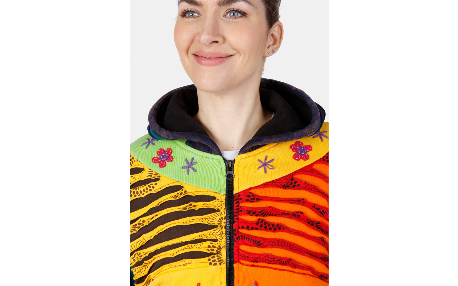 Person von Brust aufwärts mit leichtem Lächeln, trägt bunte Reißverschlussjacke mit schwarzer Kapuzeninnenfläche und gelb-grün-orangen-rot gestreiften Paneelen mit Blumen-, Stern- und wellenförmigen schwarzen Mustern, weißer Hintergrund