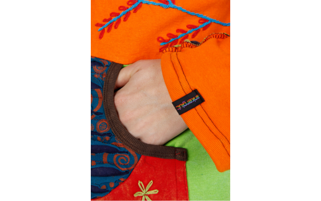 Hand steckt teilweise in der Tasche eines bunten Kleidungsstücks; oranger Ärmel mit blauer Stickerei; gemusterter dunkelblauer Stoff, rotes Feld mit gelber Stickblume; grüner Hintergrund