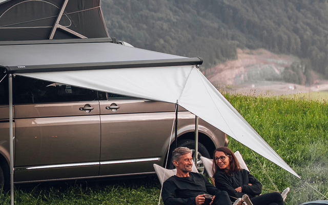 Ein Campingbus mit aufgeklapptem Dach steht auf einer Wiese, daneben sitzen zwei Personen auf Campingstühlen