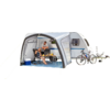 Caravane avec auvent sur l'herbe près de la plage, deux personnes assises à une table, glacière, deux vélos, plage de sable et rochers en arrière-plan, ciel bleu
