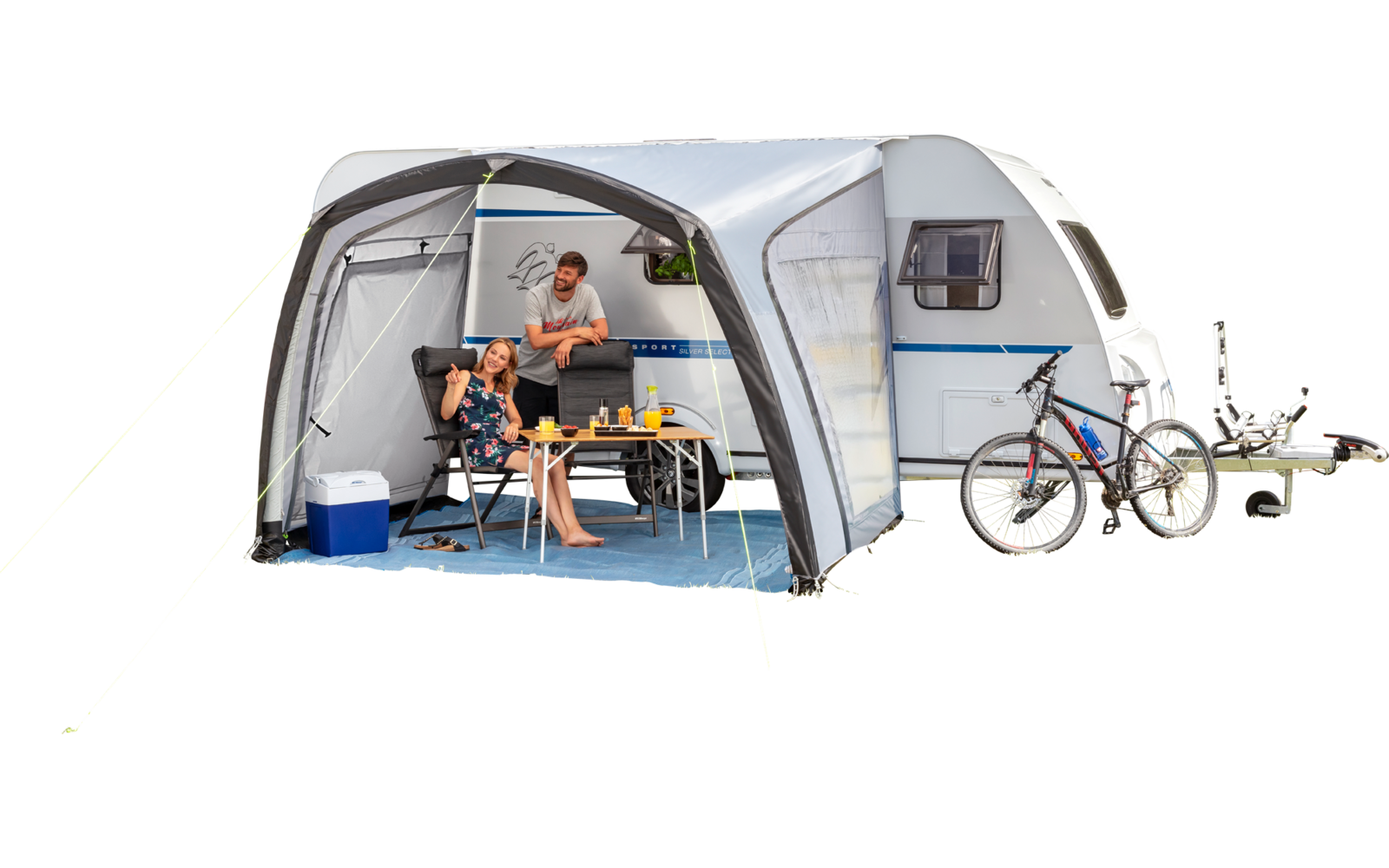 Caravane avec auvent sur l'herbe près de la plage, deux personnes assises à une table, glacière, deux vélos, plage de sable et rochers en arrière-plan, ciel bleu
