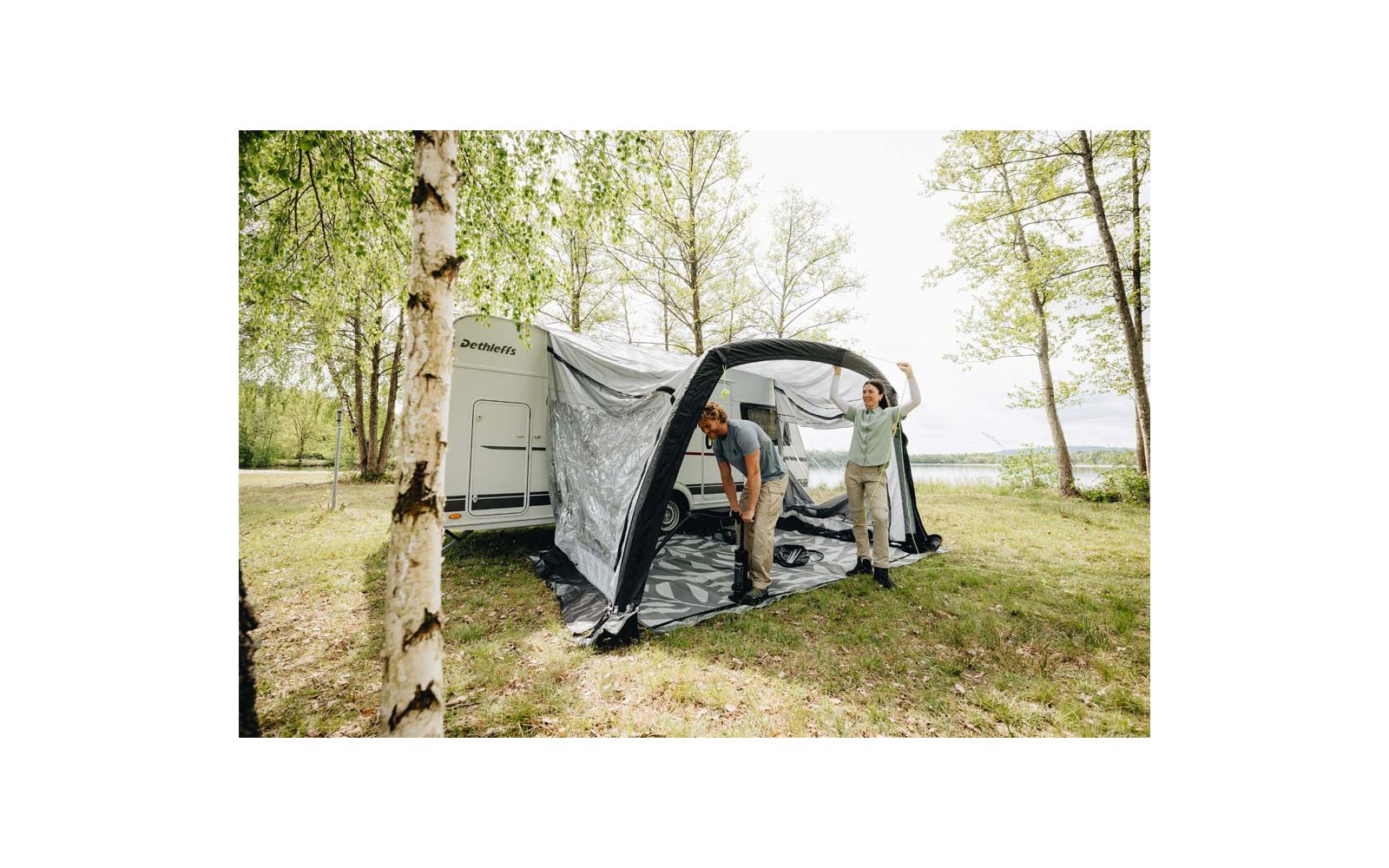 Dos personas colocan un toldo hinchable en una caravana blanca sobre césped junto a abedules y un lago