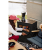 Person mit pinker Kappe kniet und öffnet die Schublade einer schwarzen Aufbewahrungsbox mit verpackten Snacks; oben steht eine Kaffeekanne; Sitzplatz mit Kissen, Holztisch mit Schalen, Fahrzeugseite, Gras und Wasser im Hintergrund