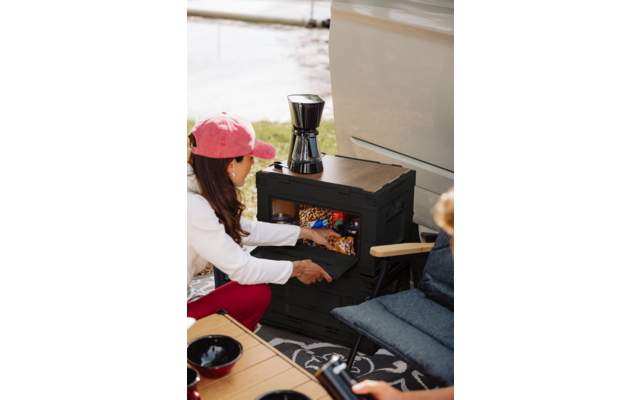Person mit pinker Kappe kniet und öffnet die Schublade einer schwarzen Aufbewahrungsbox mit verpackten Snacks; oben steht eine Kaffeekanne; Sitzplatz mit Kissen, Holztisch mit Schalen, Fahrzeugseite, Gras und Wasser im Hintergrund