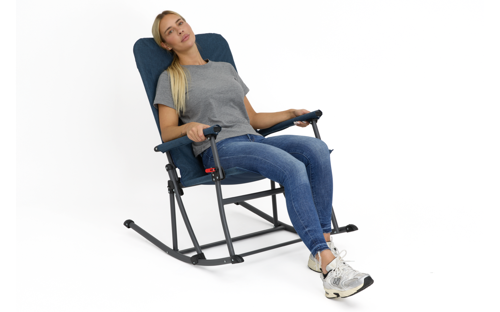Frau mit langem blondem Haar sitzt in dunkelblau gepolstertem Schaukelstuhl mit Metallgestell, trägt graues T‑Shirt, blaue Jeans und weiße Sneakers, weißer Hintergrund