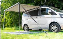 Weißer Campervan mit ausgefahrener Markise auf einer Wiese neben Bäumen