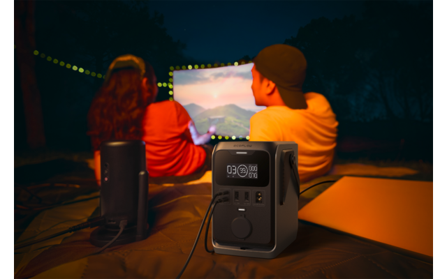 Zwei Personen von hinten sitzen im Freien vor einer beleuchteten Leinwand; im Vordergrund eine tragbare Powerstation mit Display und angeschlossenen Kabeln, zwei Lautsprecher und eine Lichterkette