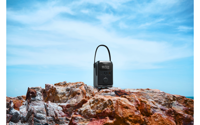 Schwarzes tragbares Gerät mit Tragegriff auf rötlich-braunen Felsen, im Hintergrund Meer und blauer Himmel mit Wolken