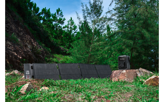 Aufgeklappte Solarpanels auf Gras neben einem Felsen, schwarze tragbare Stromstation auf dem Felsen, Nadelbäume und Hügel im Hintergrund, blauer Himmel mit Wolken