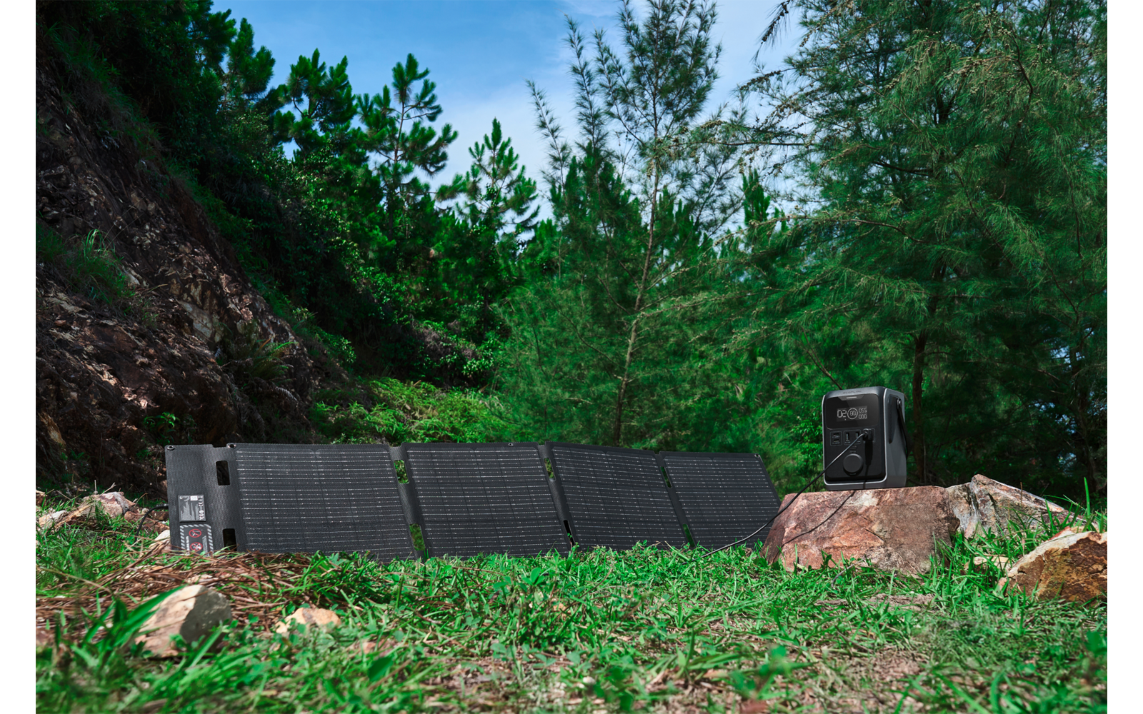 Aufgeklappte Solarpanels auf Gras neben einem Felsen, schwarze tragbare Stromstation auf dem Felsen, Nadelbäume und Hügel im Hintergrund, blauer Himmel mit Wolken