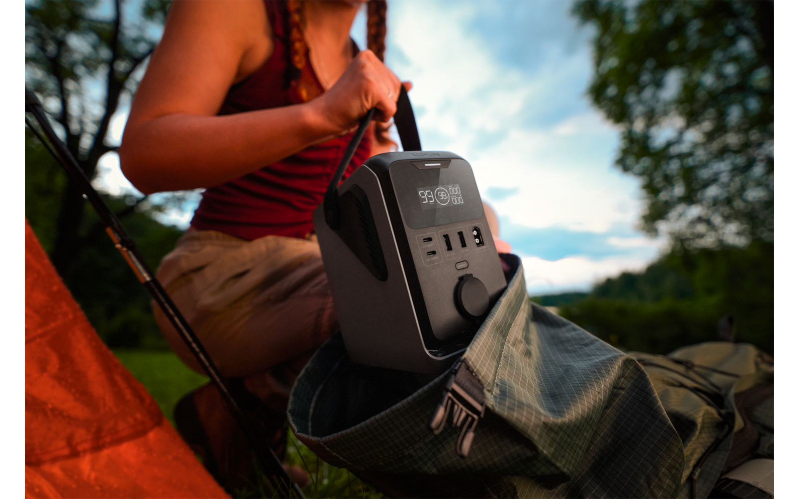 Person mit geflochtenem Haar in rotem Top setzt ein graues tragbares Stromaggregat mit Display und Anschlüssen in einen grünen Rucksack neben einem orangefarbenen Zelt, im Hintergrund Bäume und bewölkter Himmel