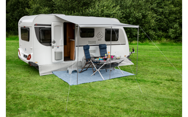 Weißer Wohnwagen mit geöffneter Tür und zwei blauen Campingstühlen neben einem Klapptisch mit Tellern und Bechern auf einer blauen Bodenmatte