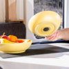 Main tenant un couvercle de bol jaune au-dessus d'un bol jaune contenant des bananes, des oranges et une pomme sur une table ; coussin et fenêtre en arrière-plan