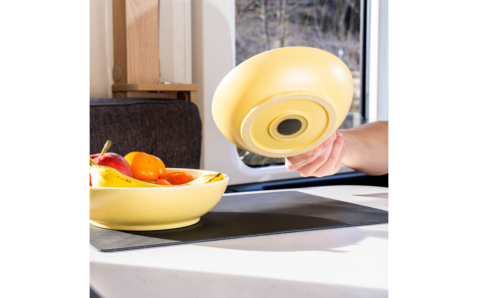 Main tenant un couvercle de bol jaune au-dessus d'un bol jaune contenant des bananes, des oranges et une pomme sur une table ; coussin et fenêtre en arrière-plan