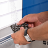 Deux mains tiennent un emballage rectangulaire noir avec texte blanc sous un jet d'eau au-dessus d'un évier en acier inoxydable ; une montre rectangulaire à bracelet noir au poignet gauche ; carrelage blanc et mur bleu en arrière-plan
