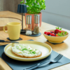 Table en bois avec une assiette contenant une tranche de pain garnie d'une pâte blanche et de fines herbes vertes hachées ; à côté une tasse jaune, un bol jaune avec des fraises, framboises et myrtilles, des salières et poivrières, un pot avec une plante verte, des couverts noirs et des sets de table noirs