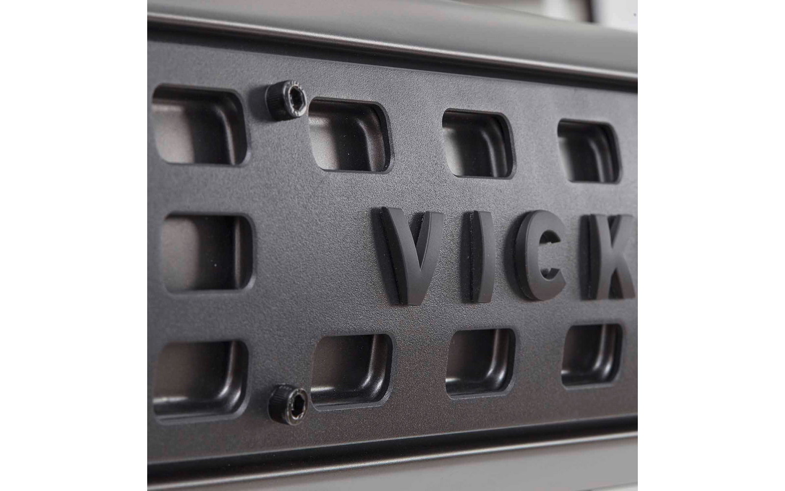 Nahaufnahme eines dunklen Metallgitters mit rechteckigen Aussparungen, erhabenen Buchstaben 'VICK' und zwei Schraubenköpfen