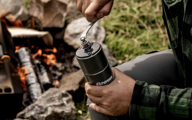 Hands of a person turning the crank of a cylindrical hand grinder, a campfire on grass in the background