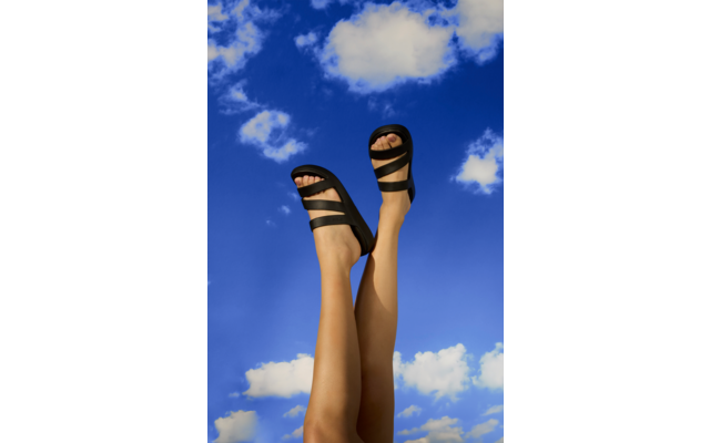 Dos piernas levantadas con sandalias negras frente a un cielo azul con nubes blancas