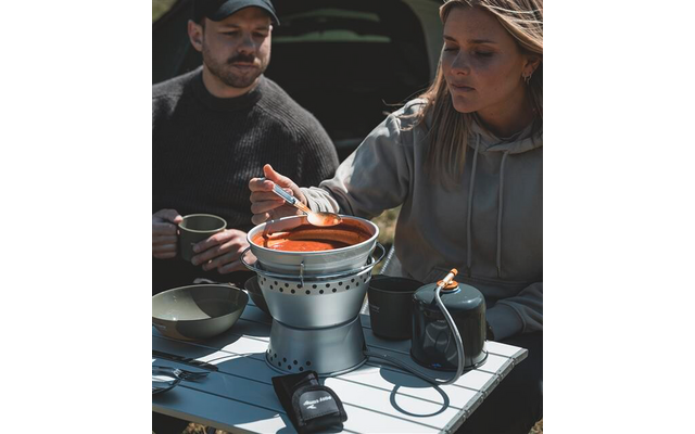 Frau rührt mit einem Löffel in einem Topf mit roter Suppe auf einem tragbaren Kocher auf einem Tisch; Mann hält eine Tasse; Schüsseln, Kanne und Besteck auf dem Tisch; geöffneter Kofferraum im Hintergrund