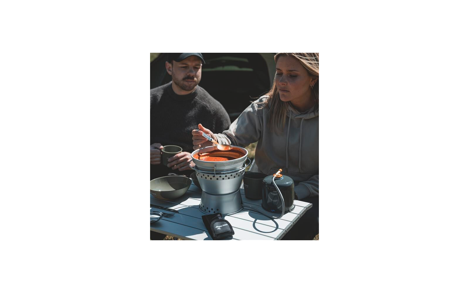 Frau rührt mit einem Löffel in einem Topf mit roter Suppe auf einem tragbaren Kocher auf einem Tisch; Mann hält eine Tasse; Schüsseln, Kanne und Besteck auf dem Tisch; geöffneter Kofferraum im Hintergrund