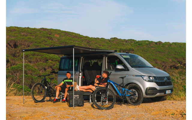 Silberner Campingvan mit ausgeklapptem Sonnendach; zwei Personen sitzen darunter, zwei Fahrräder und ein Gepäckkoffer davor; niedrige grüne Dünen und blauer Himmel im Hintergrund
