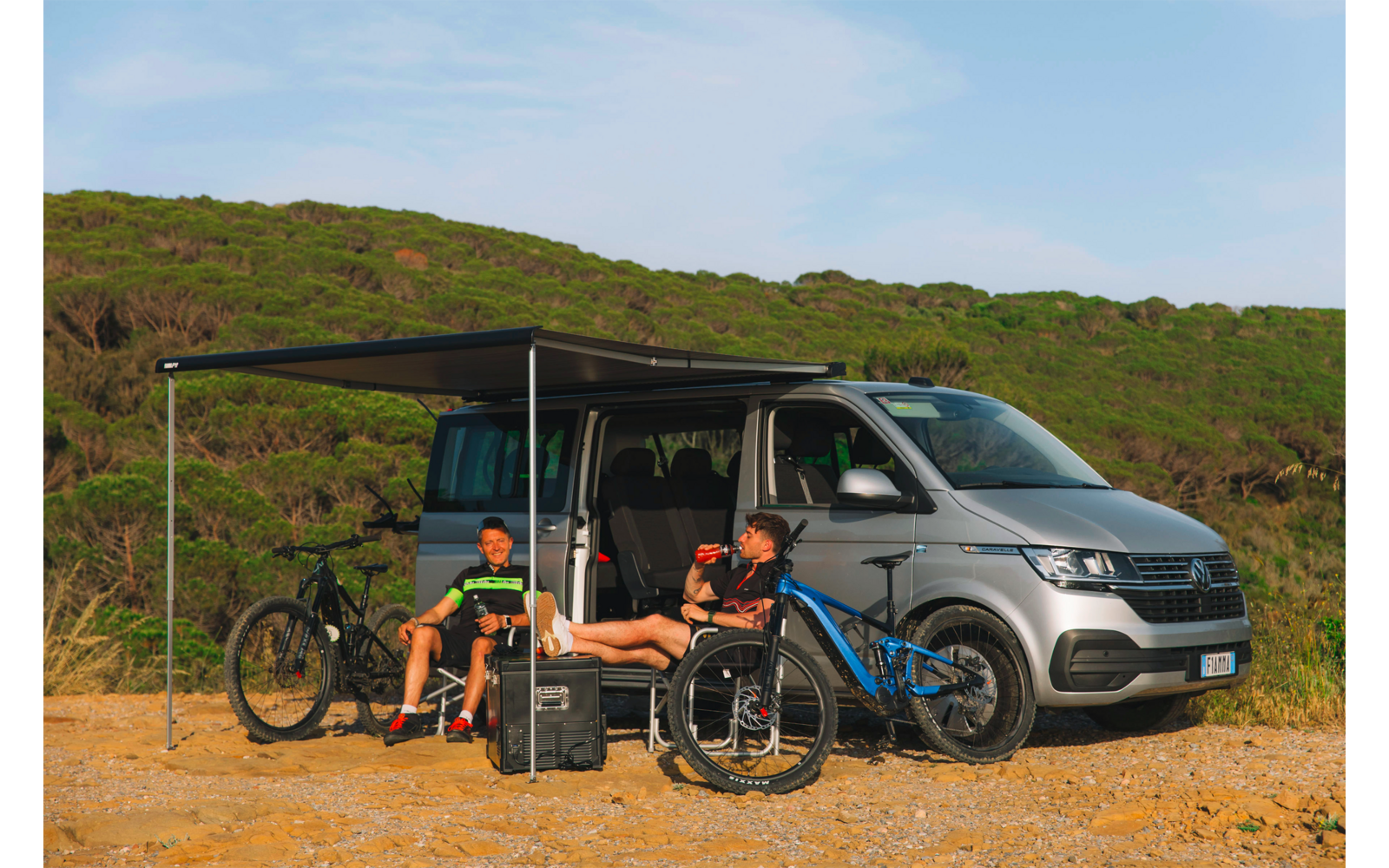 Silberner Campingvan mit ausgeklapptem Sonnendach; zwei Personen sitzen darunter, zwei Fahrräder und ein Gepäckkoffer davor; niedrige grüne Dünen und blauer Himmel im Hintergrund