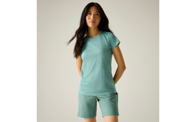 Vrouw met lang donker haar, draagt mintgroen T-shirt en groene shorts, staat tegen een effen lichte achtergrond met handen achter de rug